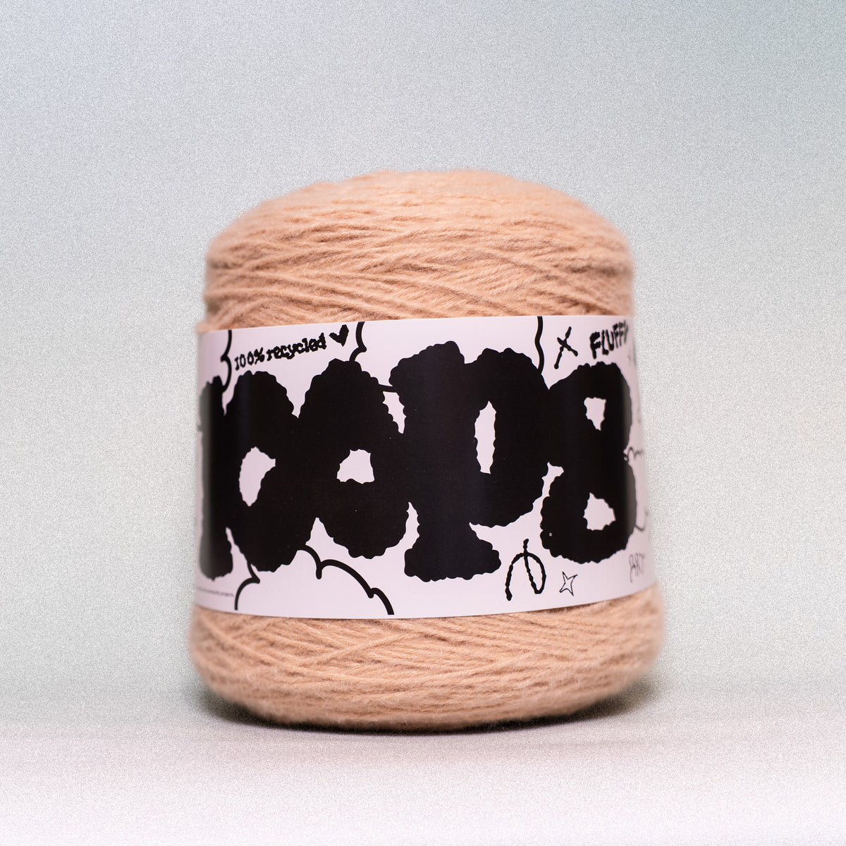 A ball of peach-colored yarn with a white paper label wrapped around it, featuring bold black graffiti-style lettering that reads 'COOL' and smaller text 'states recycled' and 'FLUFFY'. The yarn is photographed against a neutral light gray background.