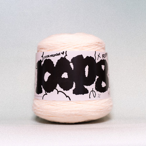 A ball of pale peach-colored yarn with a white paper label wrapped around it, featuring bold black graffiti-style lettering that reads 'COOPS'. The background is a clean, neutral light gray studio backdrop.