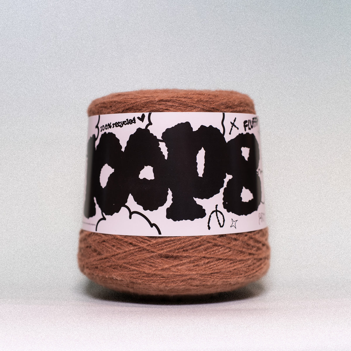 A ball of brown yarn wrapped in a white paper label with bold black text and doodles, photographed against a light gray background.