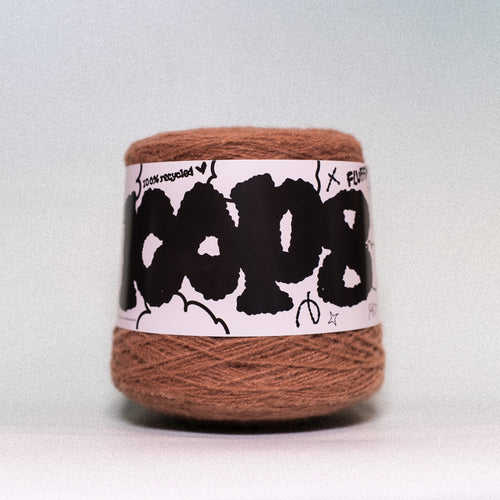 A ball of brown yarn wrapped in a white paper label with bold black text and doodles, photographed against a light gray background.