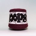 A ball of deep burgundy yarn wrapped with a white paper label featuring bold black graffiti-style lettering and small hand-drawn doodles, photographed against a neutral light gray background.