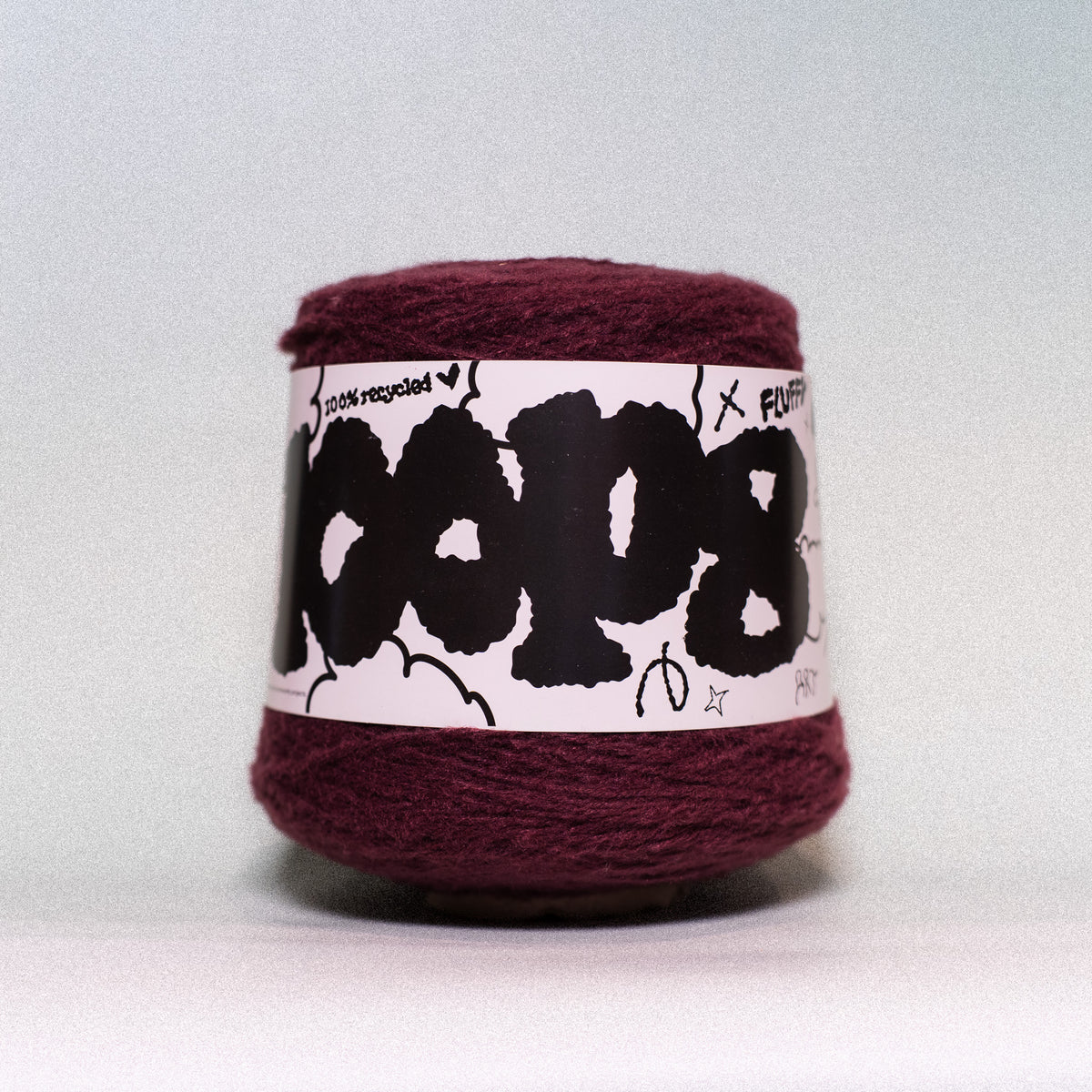 A ball of deep burgundy yarn wrapped with a white paper label featuring bold black graffiti-style lettering and small hand-drawn doodles, photographed against a neutral light gray background.