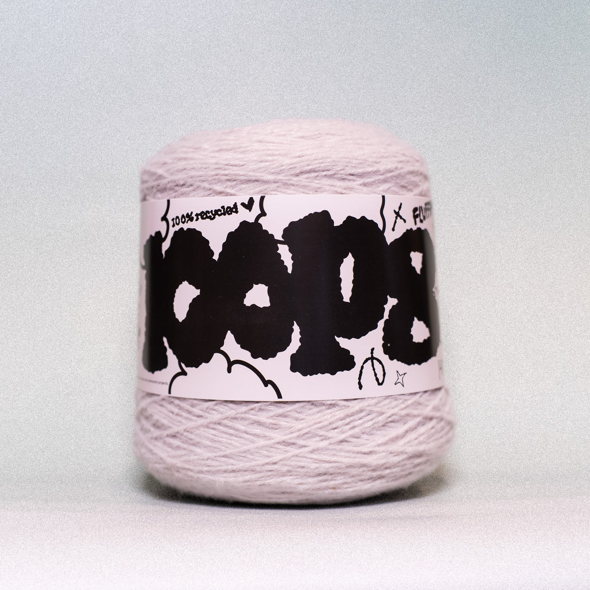 A ball of light pink yarn with a white paper label featuring bold black graffiti-style lettering, photographed against a neutral light gray background.