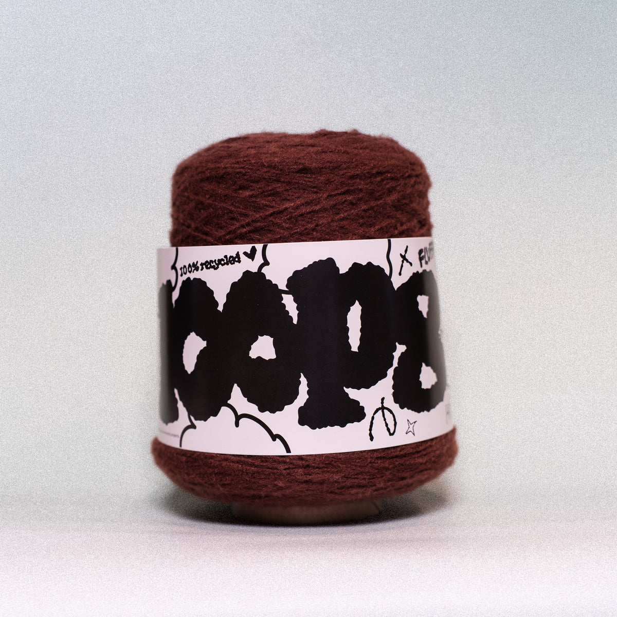 A ball of dark brown yarn wrapped in a white paper label with black graphic text, photographed against a neutral light gray background.