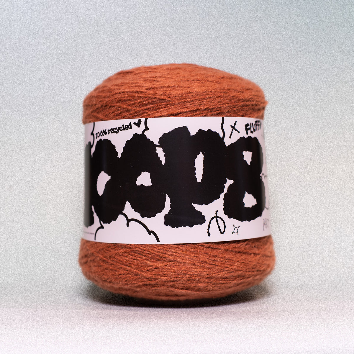 A ball of rust-orange yarn wrapped in a white paper label with bold black graffiti-style lettering, photographed against a neutral light gray background.