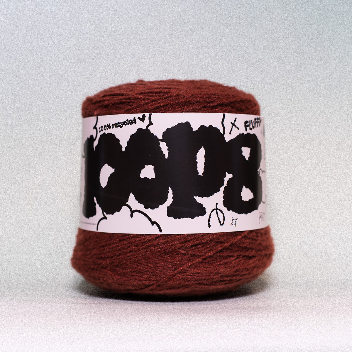 A ball of rust-brown yarn with a white paper label featuring bold black graffiti-style lettering, photographed against a neutral light gray background.
