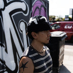 A man wearing a black knit beanie with white patches and a navy and white striped sleeveless shirt, standing outdoors in front of a graffiti-covered wall.