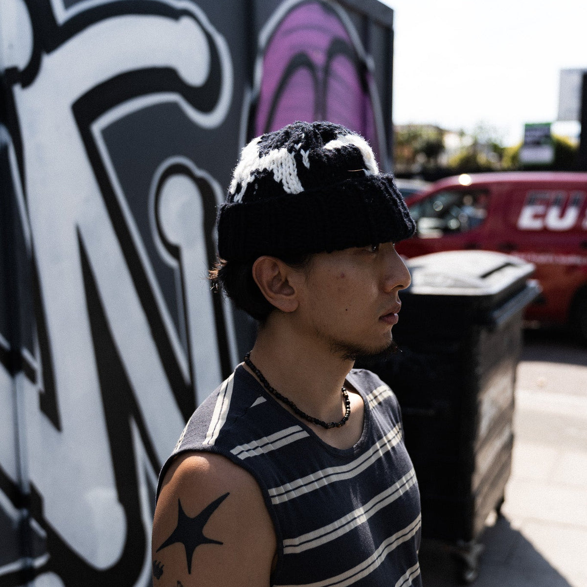 A man wearing a black knit beanie with white patches and a navy and white striped sleeveless shirt, standing outdoors in front of a graffiti-covered wall.