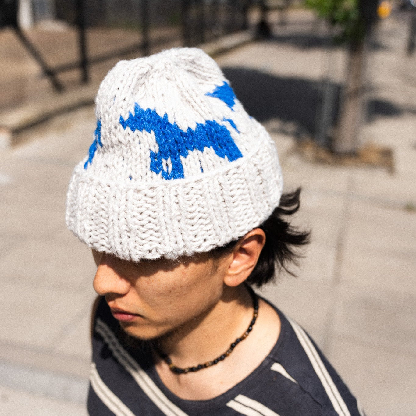 A person outdoors wearing a white knit beanie with a blue graphic pattern, paired with a navy and white striped shirt.