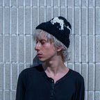 A person with light hair wearing a black knit beanie with a white graphic and a black henley shirt, standing against a gray concrete block wall.
