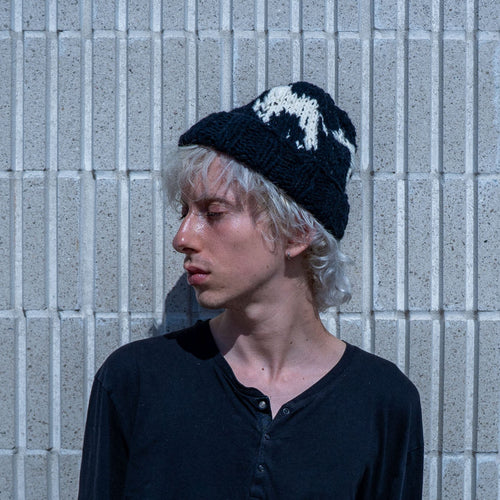A person with light hair wearing a black knit beanie with a white graphic and a black henley shirt, standing against a gray concrete block wall.