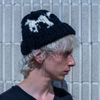A person in profile wearing a black knit beanie with a white graphic design, standing against a light gray textured wall.