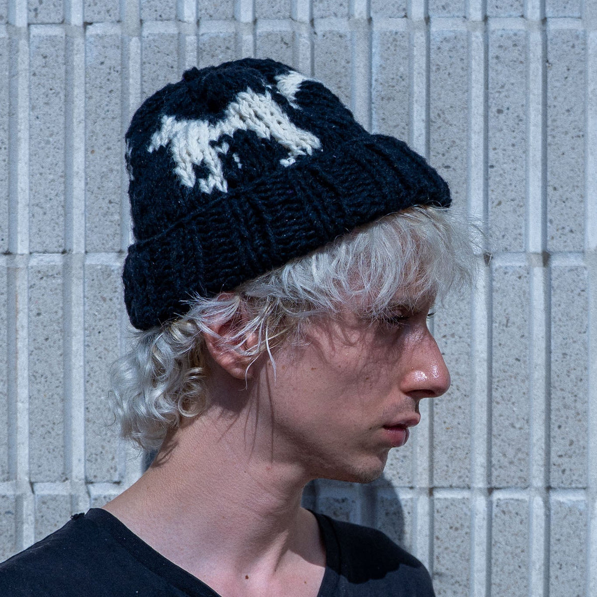 A person in profile wearing a black knit beanie with a white graphic design, standing against a light gray textured wall.