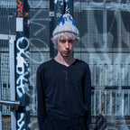 A person standing outdoors in front of a graffiti-covered metal fence, wearing a black long-sleeve henley shirt and a gray knit beanie with blue and white graphic details.