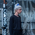 A model wearing a black long-sleeve top and black pants stands in front of a graffiti-covered metal mesh wall, wearing a gray knit beanie with blue and white graphics.
