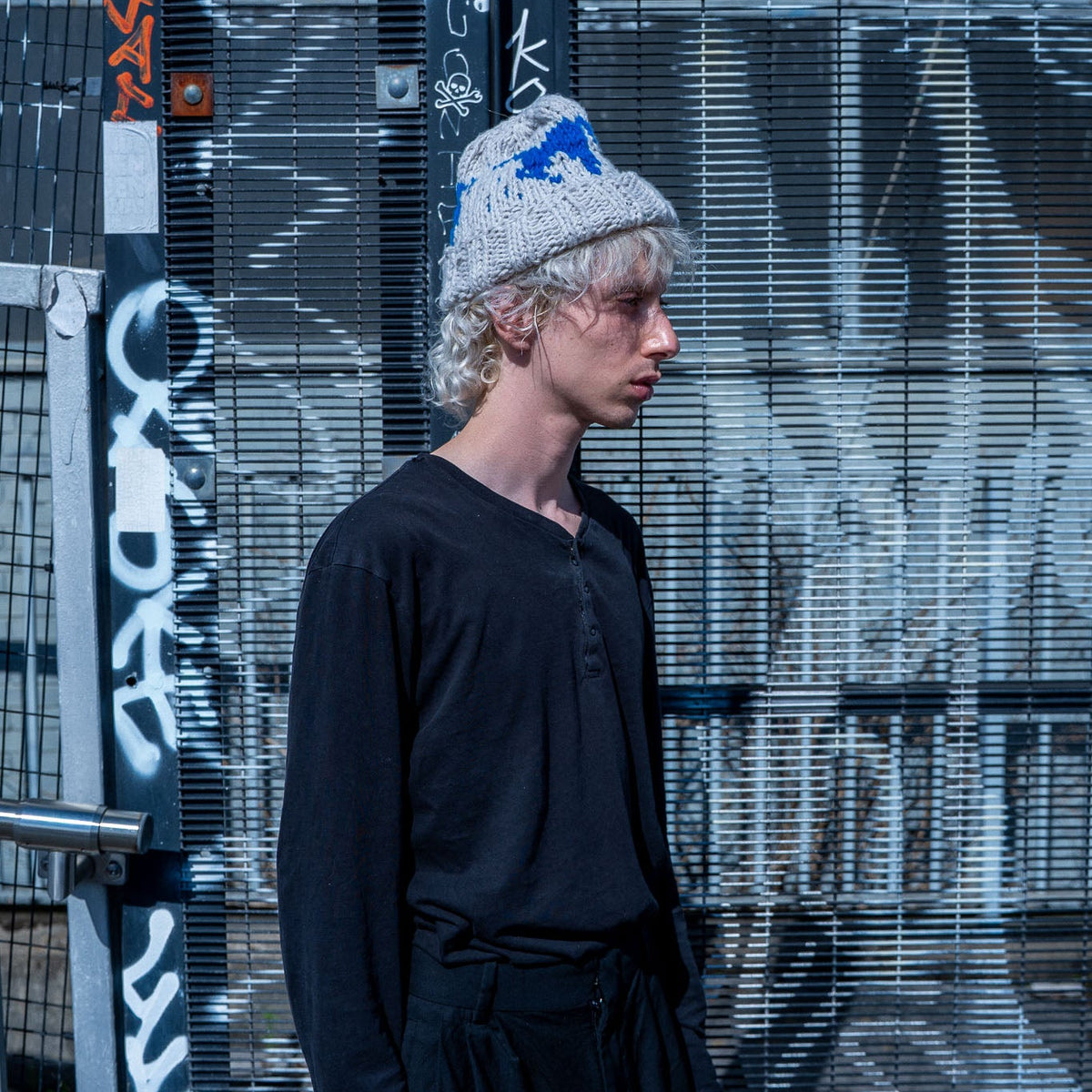 A model wearing a black long-sleeve top and black pants stands in front of a graffiti-covered metal mesh wall, wearing a gray knit beanie with blue and white graphics.