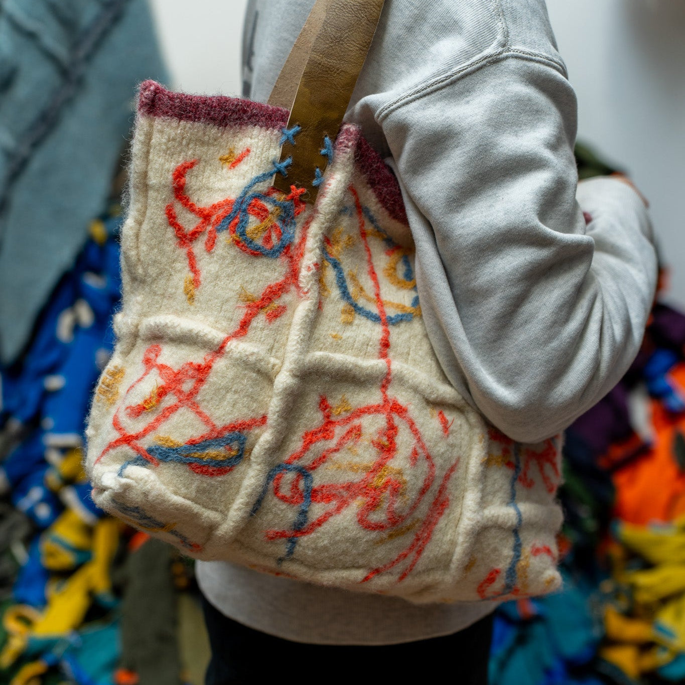 A person wearing a light gray sweatshirt carries a chunky, textured cream wool tote bag with colorful abstract yarn-like embroidery in red, blue, and orange, featuring a brown shoulder strap.