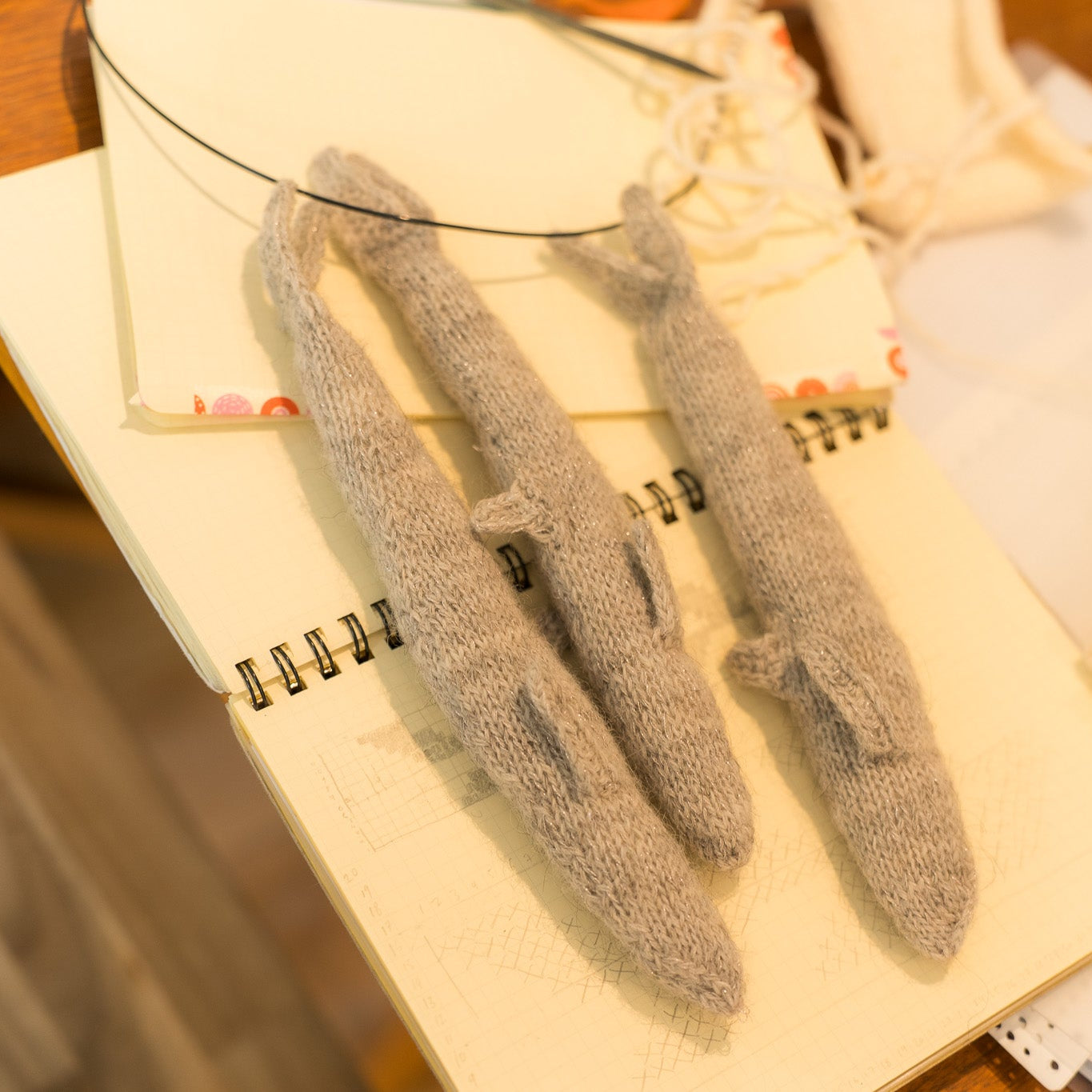 Three gray knitted fish-shaped accessories laid on an open cream notebook on a wooden table, with additional cream fabric pieces nearby.