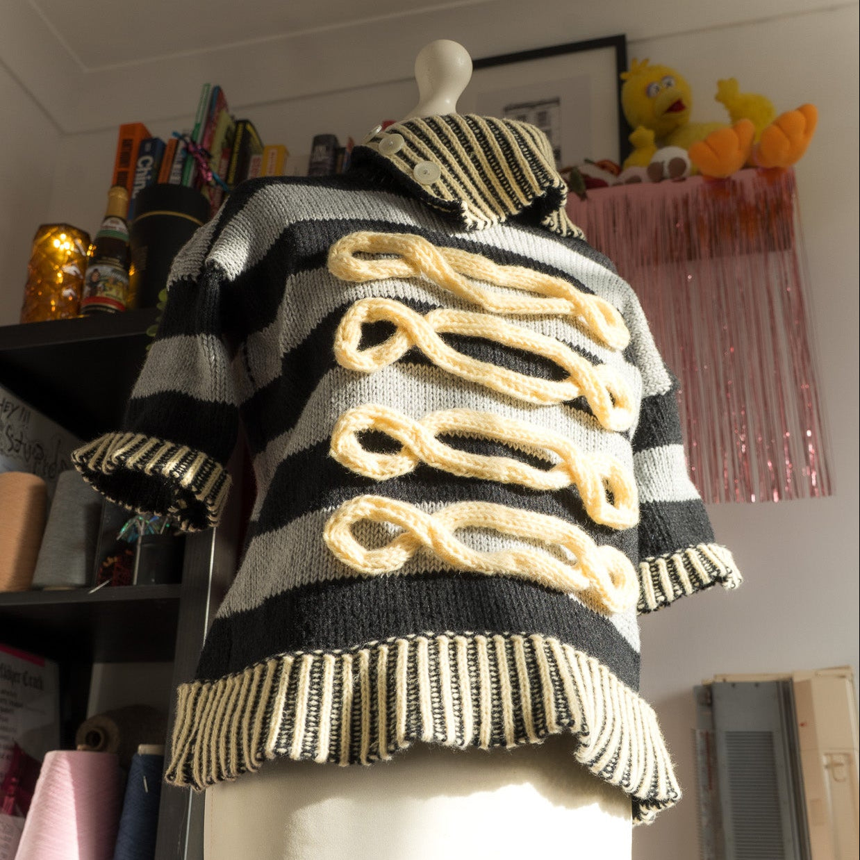 A black and white striped knit sweater with beige rope-like decorative details down the front, displayed on a white mannequin torso in a room with shelves and toys in the background.