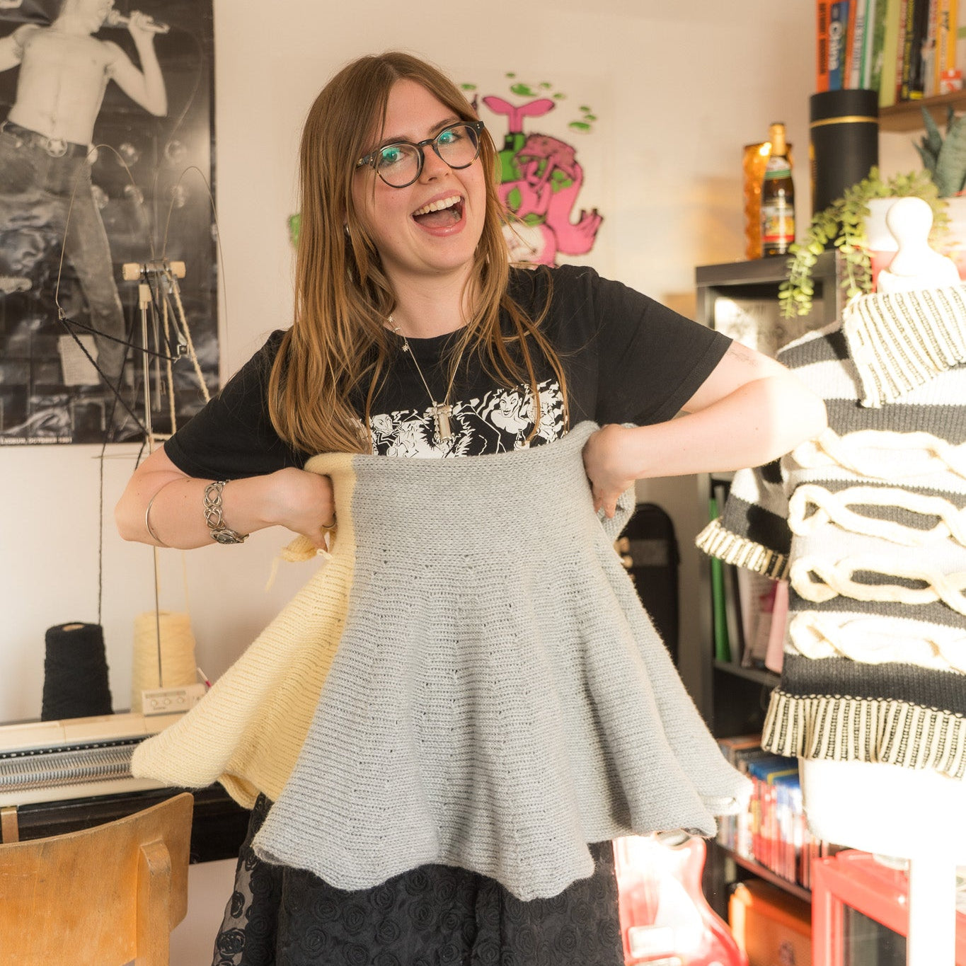 A woman with glasses and a black graphic t-shirt holds up a light gray knitted flared cape or poncho in a cluttered room. She is also holding a yellow garment in her other hand.